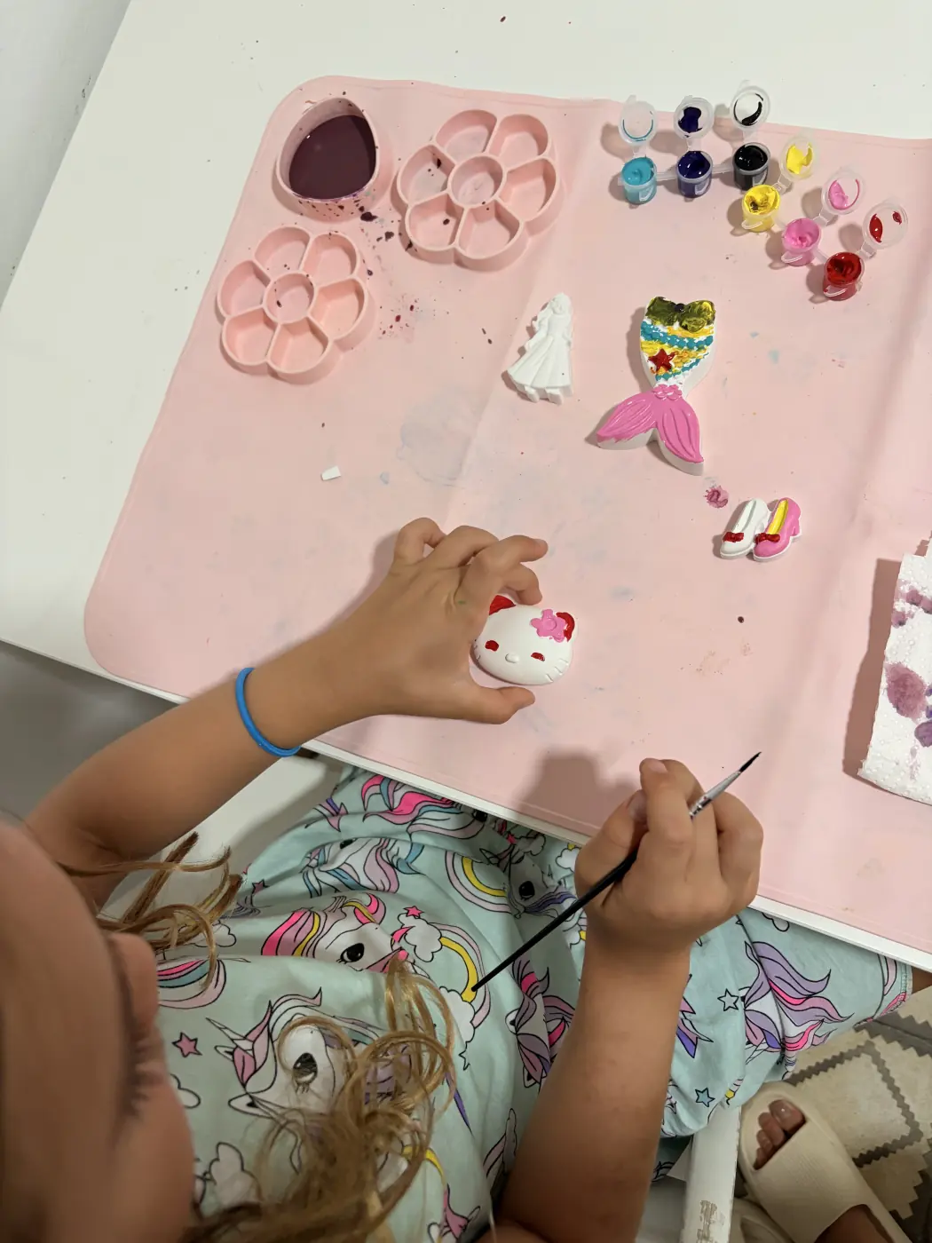 A child paints plaster figures at a pink craft table, with a mermaid, princess, tiny shoes and paint pots nearby.