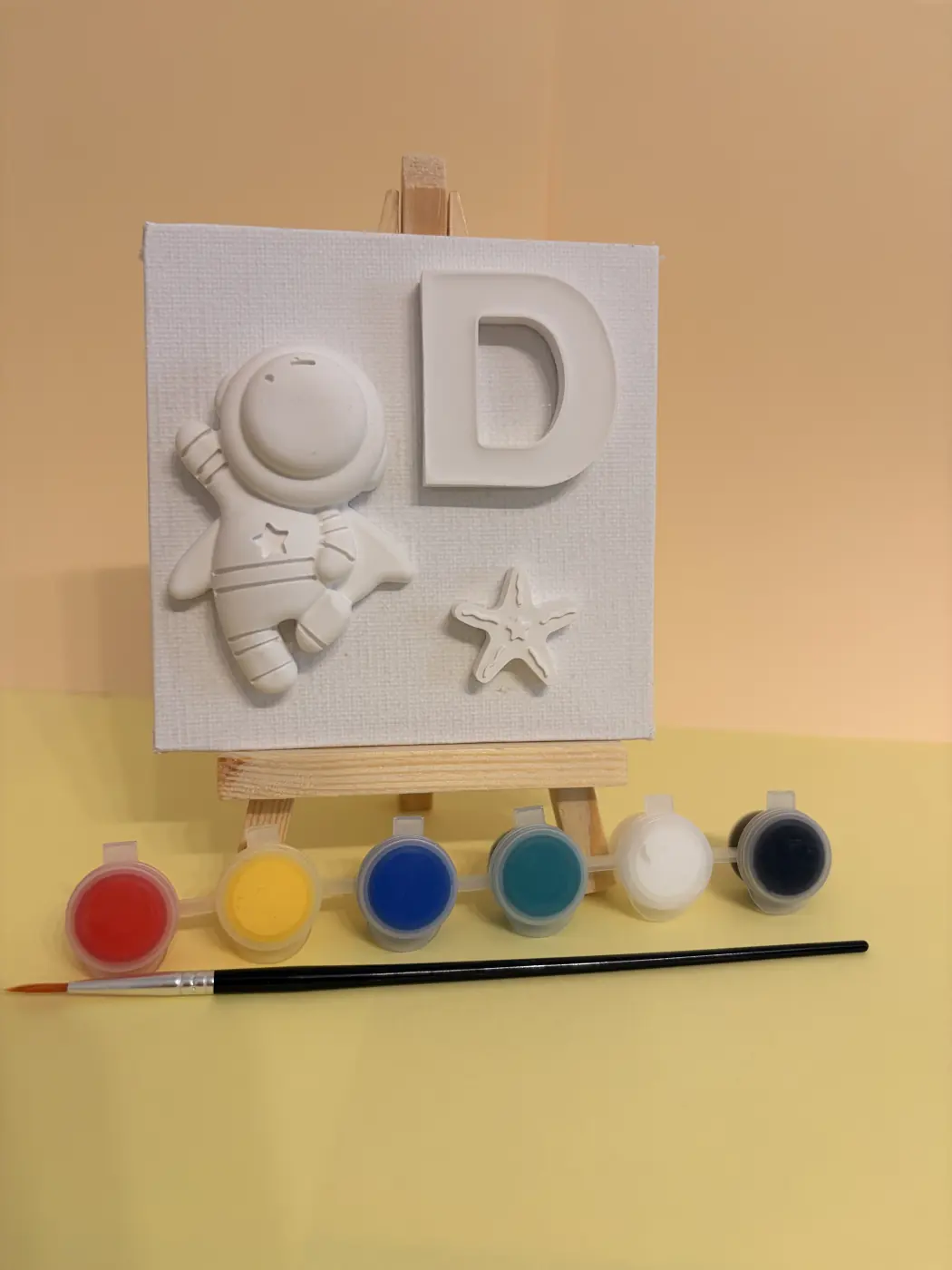 Unpainted astronaut, letter D and star arranged on a small canvas with a mini easel, paints and brush.