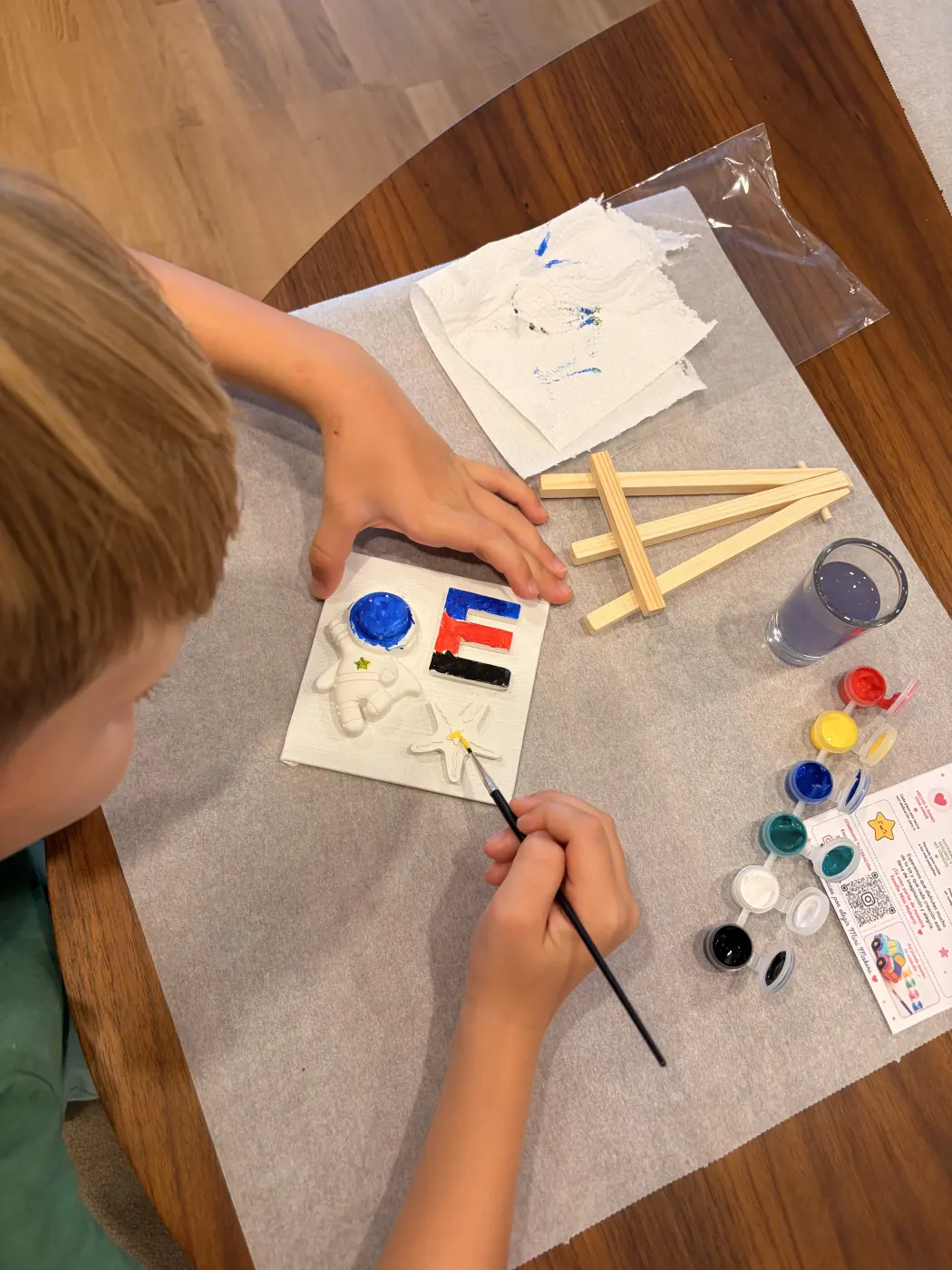 A child paints an astronaut, letter E and star on a small canvas, with paints and mini easel pieces nearby.