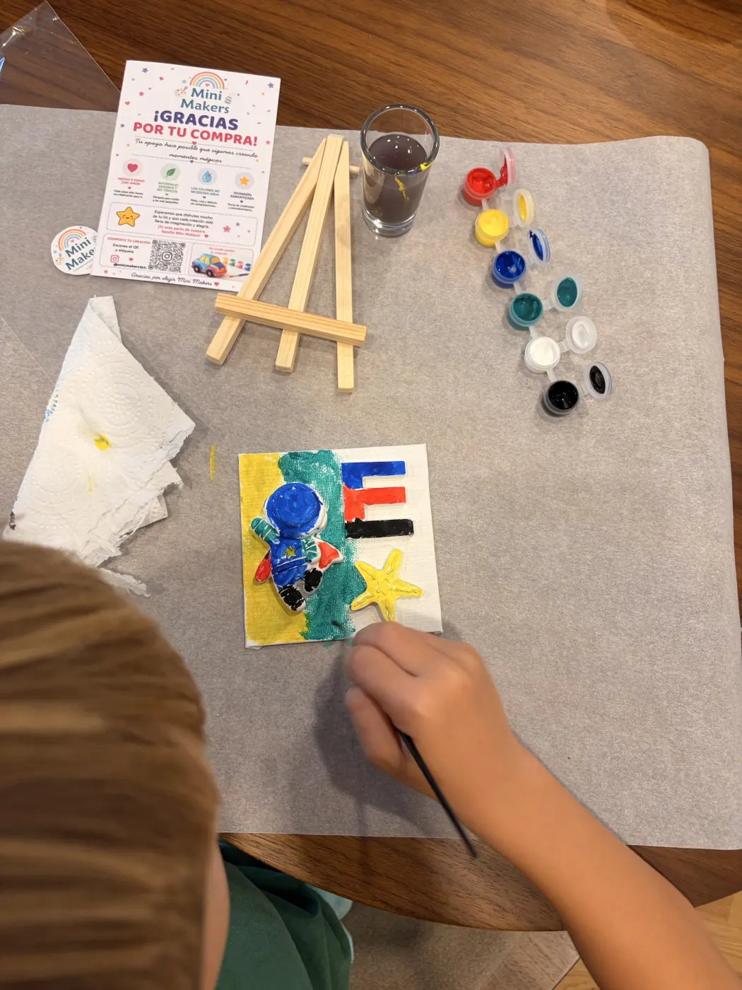 A child paints a blue astronaut and a colourful letter E on a small canvas, surrounded by paints and a mini easel.