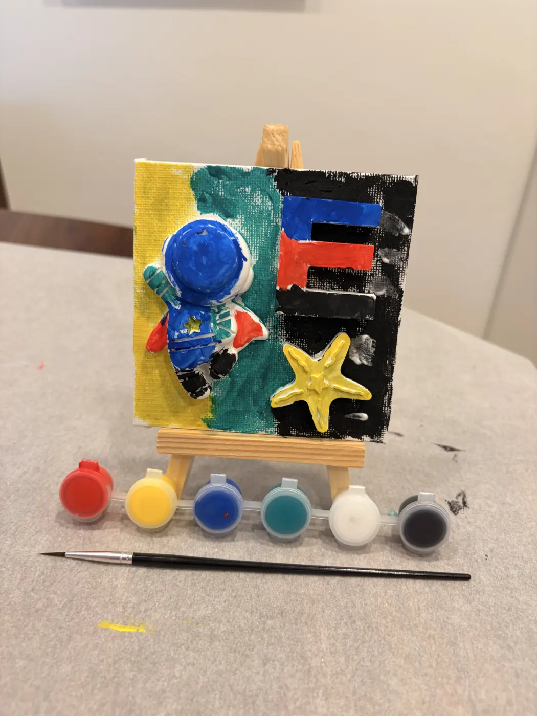 Finished artwork of a blue astronaut beside a colourful letter E and yellow star, displayed on a mini easel with paints and a brush.
