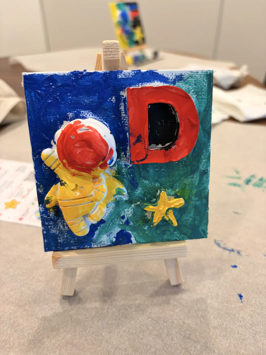 Finished artwork of a yellow astronaut beside a red letter D and yellow star, displayed on a mini easel.